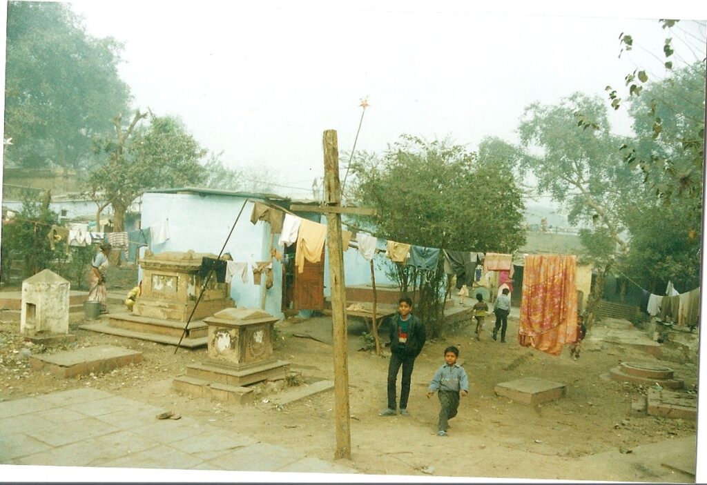 Washing line strung between tall wooden cross and grave stone. drying laundry. Resident children walking between graves.