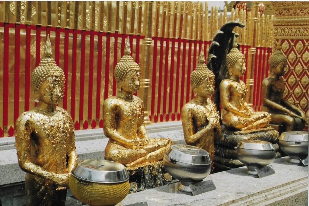 Thailand travel writing. Four gold leaf buddhas sit cross legged side by side in front of a Chiang Mai temple wall.  