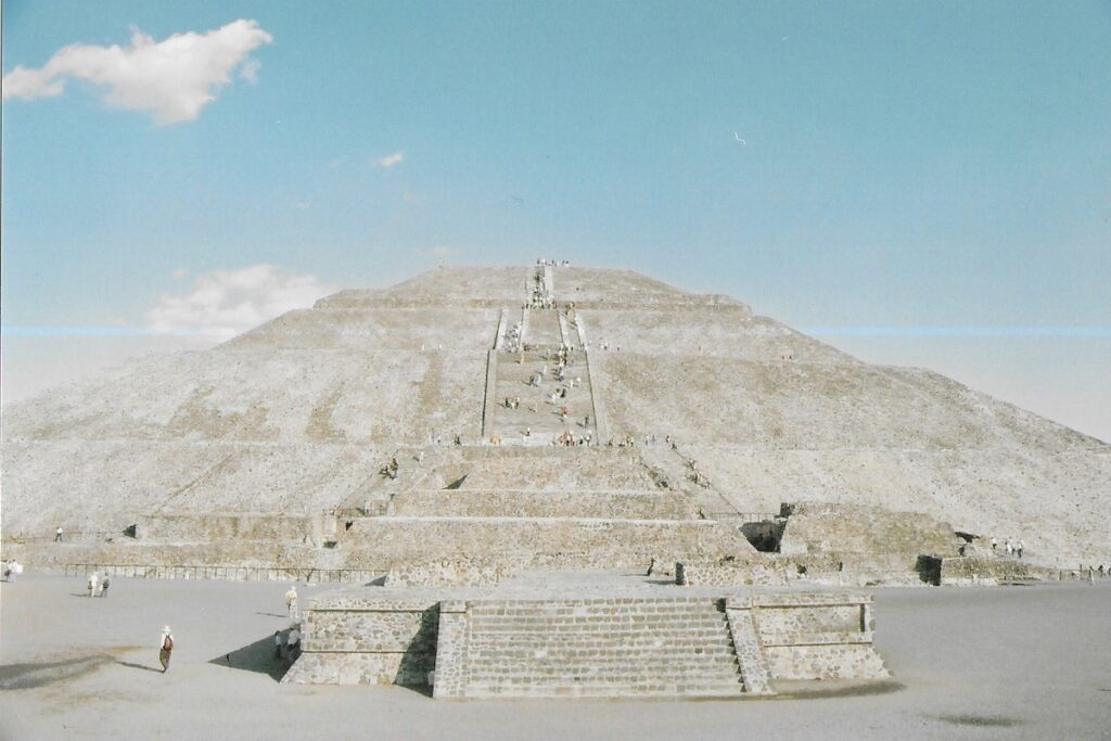 Huge Aztec stepped pyramid rises high into a sun baked sky. People climbing up the steps are reduced to the size of ants.