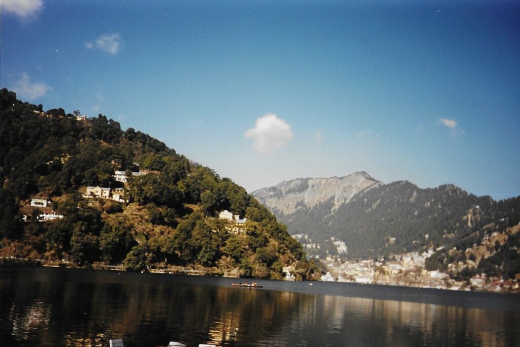 Nainital lake Nainital's serene lake glistens. A lone boat sits in the middle. Mountain's and hills rise in the background, with houses built into their sides.