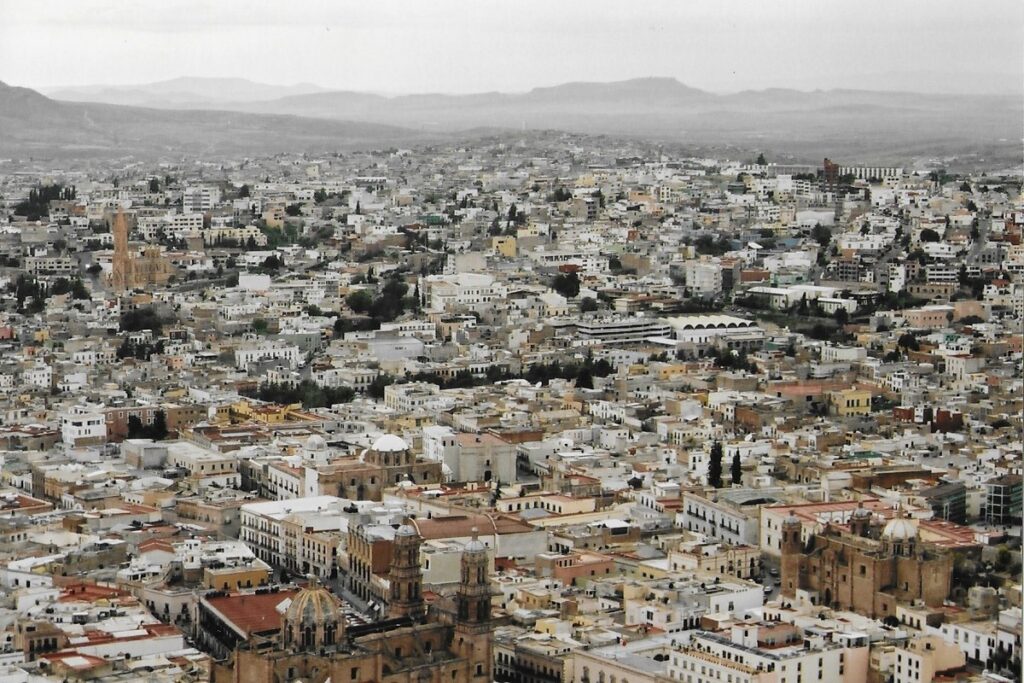 A panoramic bird's eye view of Zacatecas city taken from high up a hill top. Through the distant haze are mountain ridges. 